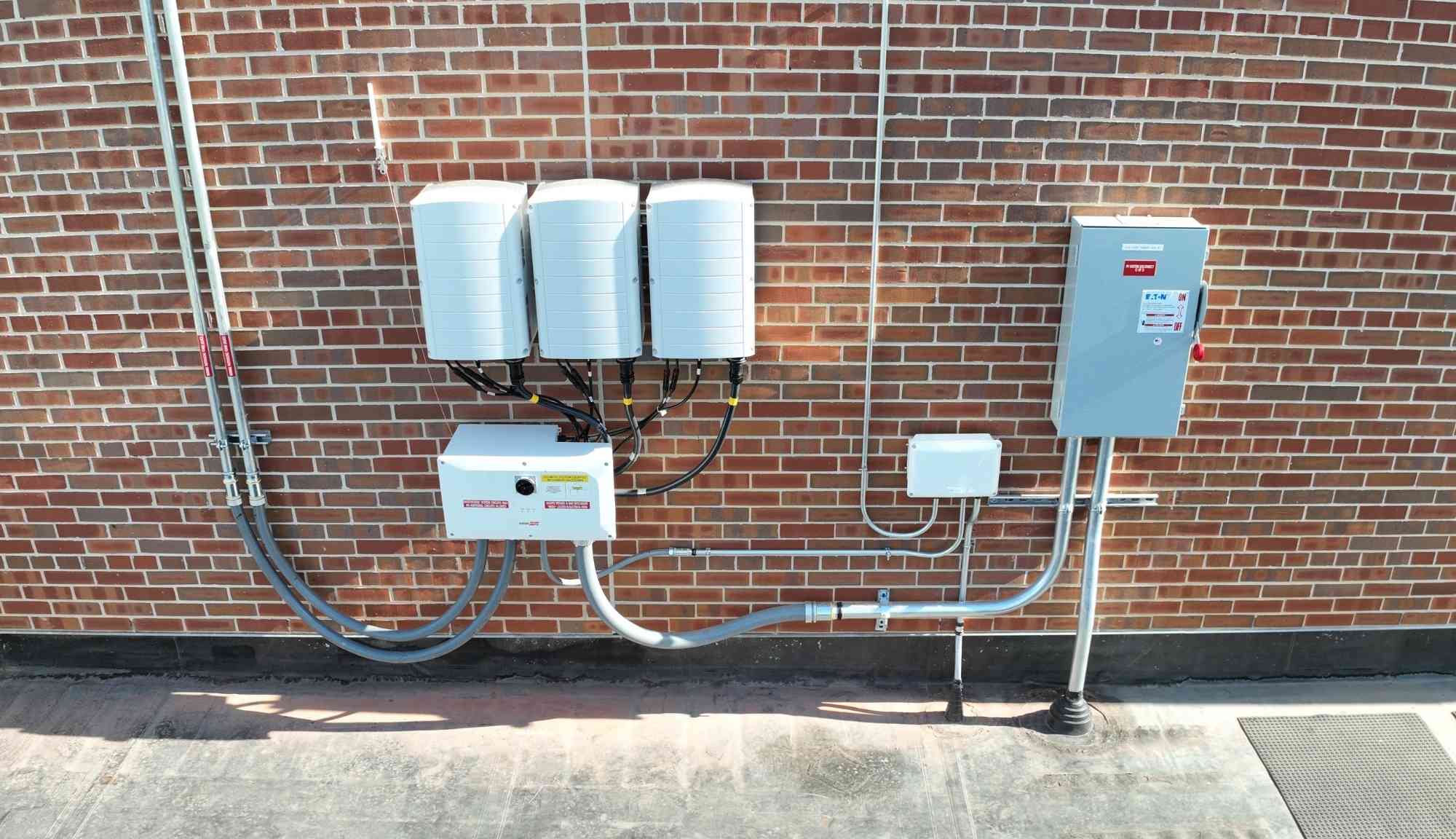 Wall mounted solar inverters and electrical equipment connected to a rooftop solar array on a brick building.