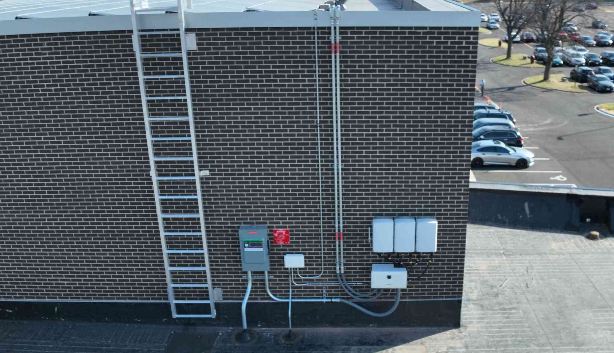 Exterior wall view showing rooftop solar electrical equipment, conduit runs, and access ladder on a commercial building.
