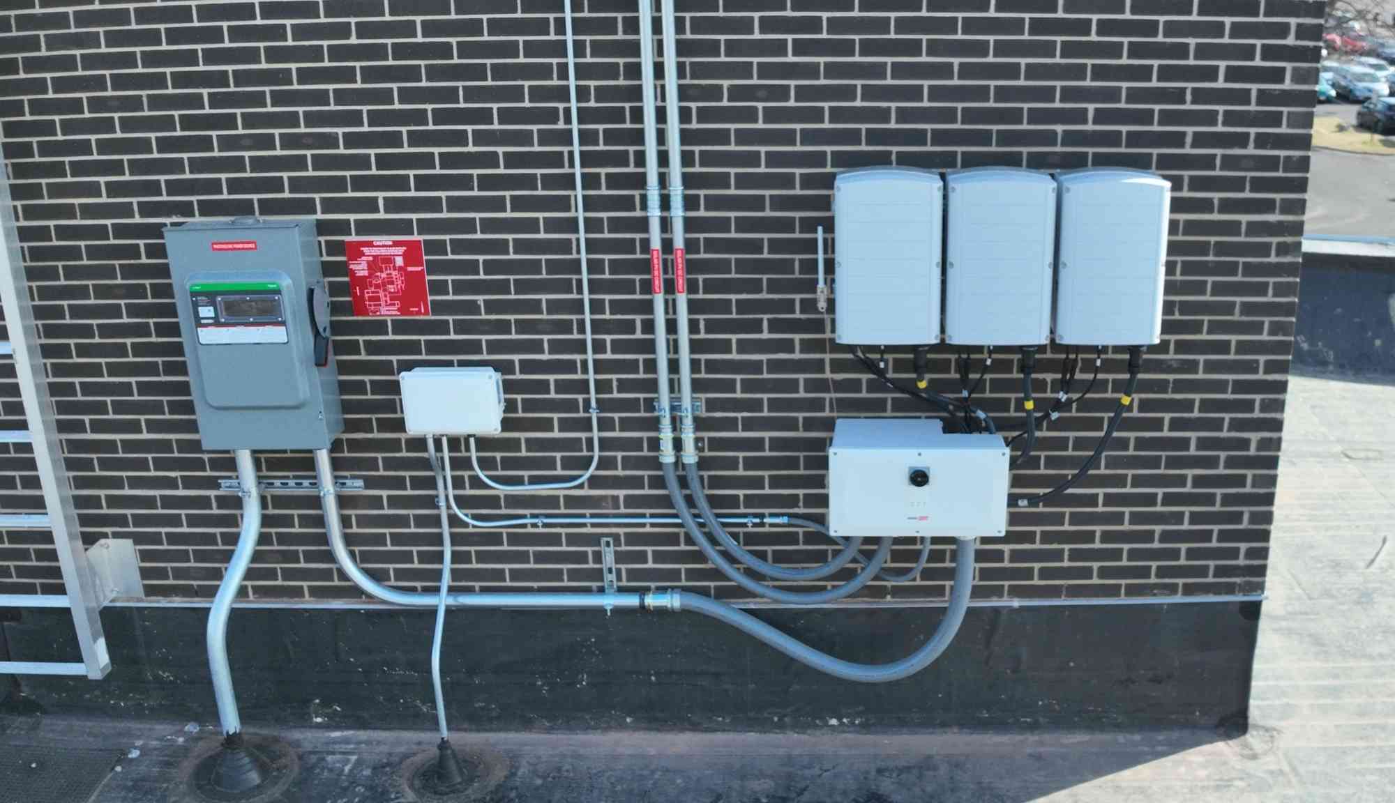 Wall mounted solar inverters, disconnects, and electrical conduit installed on a brick commercial building.