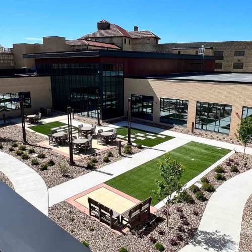 Exterior view of a modern courtyard with landscaped walkways, seating areas, and large windows on an institutional building.